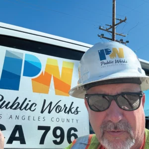 LA County Public Workes member Jimmy Dollison is in front of a County truck. He's wearing sunglasses and is wearing a white safety helmet with the Public Works logo.